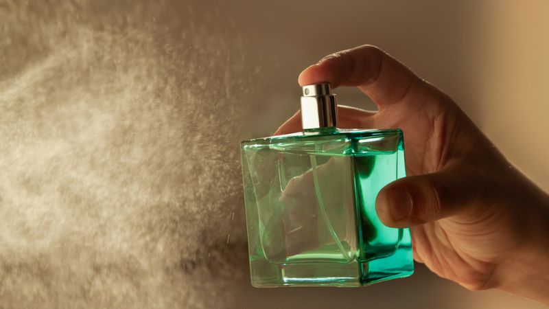 Imagem de um perfume em frasco verde sendo pressionado com as gotículas do produto sendo representadas em foto.