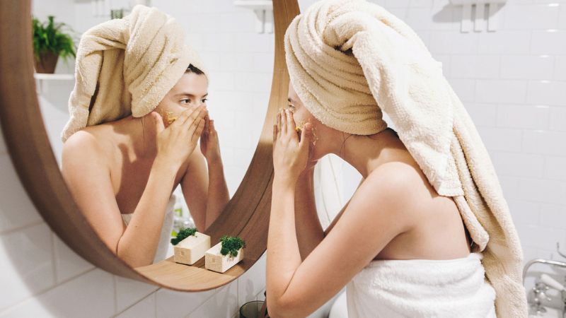 mulher na frente do espelho e com uma toalha branca na cabeça esfoliando o rosto.